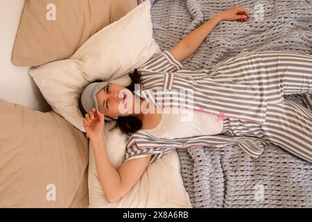 Frau in grauem gestreiftem Pyjama und Schlafmaske lachend im Bett Stockfoto