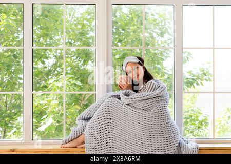 Entspannte Frau in gestreiftem Pyjama, die am Fenster mit Becher und Decke sitzt. Stockfoto