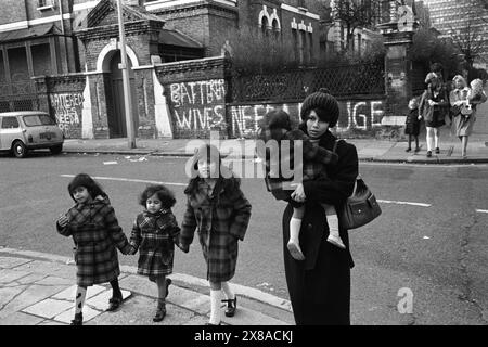 Chiswick Women's Aid 1970s UK. Eine Gruppe von verprügelten Frauen aus dem Schutzgebiet und asiatische Mutter mit ihren drei Kindern laufen zu lokalen Geschäften. An der Wand schrieb: „Bettered Wives Need Refuge“ Richmond, London, England November 1975. HOMER SYKES Stockfoto