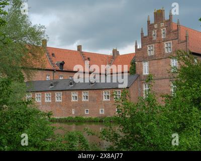 Ein imposantes Backsteingebäude mit einem roten Dach, mehreren Fenstern, umgeben von grüner Vegetation und einem Fluss unter einem bewölkten Himmel, rote Backsteinburg mit Roun Stockfoto