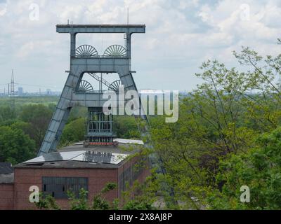 Großer gewundener Turm über einem alten Industriebau, umgeben von grüner Landschaft und unter einem bewölkten Himmel, Bergbauturm einer stillgelegten Zeche in der Stockfoto