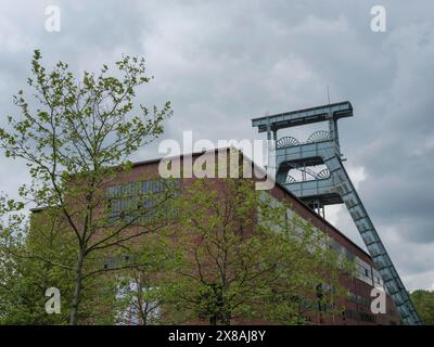 Industriebau mit gewundenem Turm und grünen Bäumen im Vordergrund unter bewölktem Himmel, gewundener Turm einer stillgelegten Zeche im grünen Ruhrgebiet Stockfoto