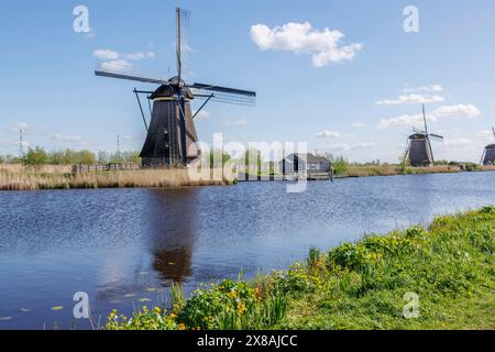 Eine Windmühle am Ufer eines Flusses neben einer kleinen Hütte unter blauem Himmel mit Wolken, umgeben von Gras und Natur, Windmühlen auf einem Fluss im Nether Stockfoto