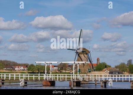 Historische Windmühle neben einer Brücke über einen ruhigen Fluss an einem bewölkten Tag, alte Windmühlen an einem Fluss in den Niederlanden inmitten von weiten Feldern dagegen Stockfoto