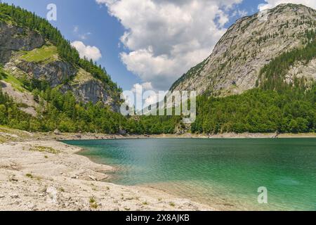 Smaragdgrüner Bergsee umgeben von steilen, bewaldeten Berghängen und felsigen Klippen, unter teilweise bewölktem Himmel, glitzernder grüner Bergsee Stockfoto