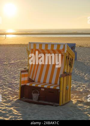 Einzelne, orange und weiß gestreifte Liege bei Sonnenuntergang am Strand, Sonnenuntergang am Meer mit vielen, bunten Liegen am Strand, warmes Licht Atmo Stockfoto