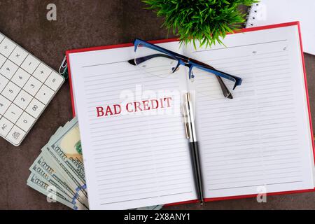 Text „schlechte Gutschrift“ auf der offenen Seite des Business-Notizbuchs neben einer Brille, einem Taschenrechner, Dollar, einer Pflanze und einem Stift. Stockfoto