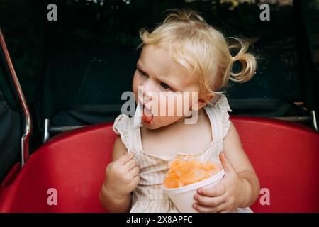 Ein Kleinkind isst orange rasiertes Eis in einem roten Wagen Stockfoto