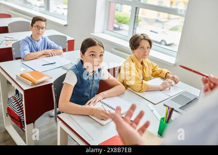 Eine Gruppe von Kindern hört aufmerksam einem männlichen Lehrer zu, während sie an Schreibtischen in einer hellen, lebendigen Klassenzimmer-Umgebung sitzen. Stockfoto