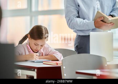 Ein junges Mädchen sitzt an ihrem Schreibtisch, vertieft in ein Buch und benutzt einen Taschenrechner. Das Klassenzimmer ist hell und lebendig, mit einem Lehrer, der sie unterrichtet Stockfoto