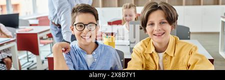 Zwei Jungen sind tief konzentriert, sitzen an einem Tisch in einem lebhaften Klassenzimmer, während ihr Lehrer die Gruppe der Schüler unterrichtet. Stockfoto