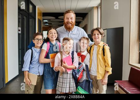 Ein engagierter Lehrer steht in einer multikulturellen Gruppe von Kindern in einem lebendigen Schulflur und engagiert sie in einer interaktiven Lerneinheit. Stockfoto