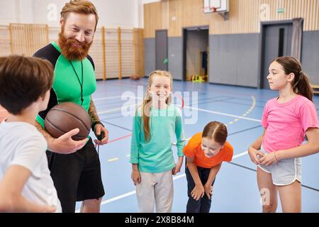 Ein Lehrer hält einen Basketball, während eine Gruppe von Kindern in einem hellen, lebhaften Klassenzimmer um ihn herum steht. Stockfoto