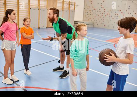 Eine Gruppe von Leuten, angeführt von einem männlichen Lehrer, die in einem Fitnessstudio herumstehen und Basketball halten, nahmen an einer Basketballstunde Teil. Stockfoto