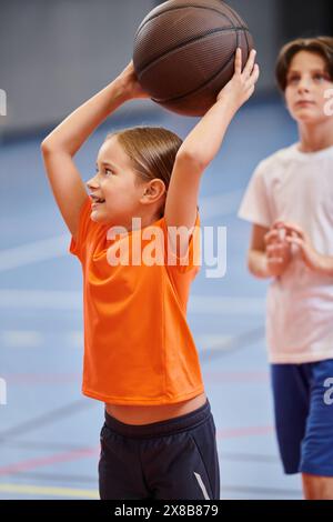 Ein junges Mädchen hält mit Freude einen Basketball hoch oben in der Luft und strahlt ein Gefühl der Begeisterung und Leidenschaft für das Spiel aus. Stockfoto