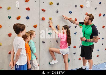 Ein männlicher Lehrer hilft einer vielfältigen Gruppe von Kindern, während sie lernen, auf eine bunten Kletterwand in einer lebendigen Klassenzimmer-Umgebung zu klettern. Stockfoto