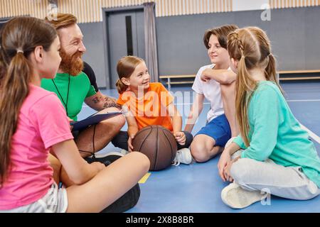 Eine Gruppe von verschiedenen Kindern sitzt auf dem Boden und hört den Anweisungen eines männlichen Lehrers zu, ein Basketball in der Mitte. Stockfoto