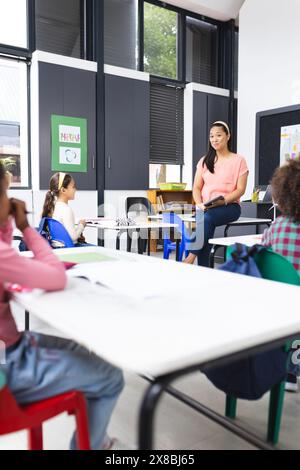 In der Schule sitzt eine junge, birassische Lehrerin auf einem Schreibtisch gegenüber ihren verschiedenen Schülern Stockfoto