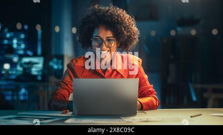 Schöne multiethnische brasilianische Frau, die spät in der Nacht auf Laptop-Computer arbeitet. Junger Creative Manager, der Spaß im Büro hat, mit Freunden plaudert, lacht und glücklich ist. Stockfoto