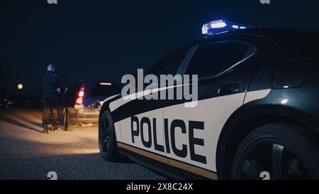 Porträt eines kaukasischen Polizisten mittleren Alters, der sich mit Vorsicht einem angezogenen Fahrzeug nähert. Ein betrunkener Fahrer wird von einem Offizier angehalten, um seine Papiere zu überprüfen. Kamerafokus auf Polizeiauto Stockfoto