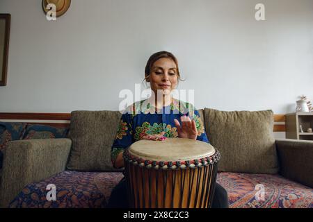 Weitwinkel-Vorderansicht einer erwachsenen lateinischen Frau, die zu Hause glücklich mit einer Djembe-Trommel sitzt. Sie trägt ein Hemd im afrikanischen Stil und konzentriert sich auf das Spiel Stockfoto