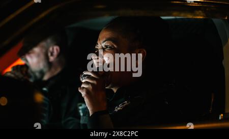 Porträt von zwei Polizisten, die im Auto sitzen und nachts über die Straßen schauen. Weibliche schwarze Polizistin beißt auf einen Donut, während sie auf die Disposition wartet. Polizist-Partner machen eine Pause Stockfoto