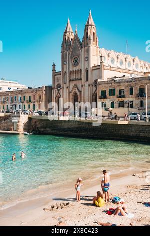 Die Menschen baden im flachen türkisfarbenen Wasser am Balluta Bay Beach in St. Julian's, Malta. Die neogotische Karmeliterkirche ist dahinter zu sehen Stockfoto