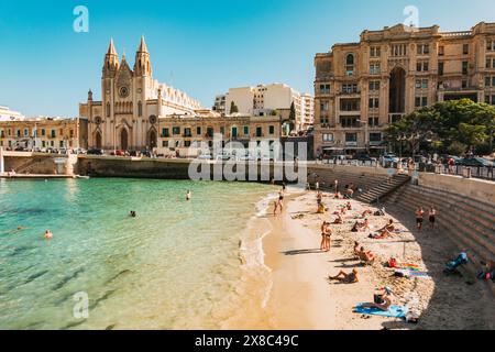 Die Menschen baden im flachen türkisfarbenen Wasser am Balluta Bay Beach in St. Julian's, Malta. Die neogotische Karmeliterkirche ist dahinter zu sehen Stockfoto