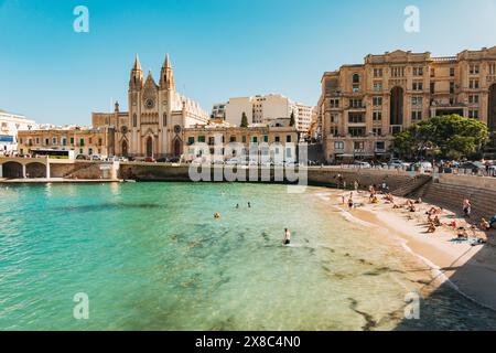 Die Menschen baden im flachen türkisfarbenen Wasser am Balluta Bay Beach in St. Julian's, Malta. Die neogotische Karmeliterkirche ist dahinter zu sehen Stockfoto