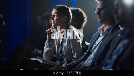 Junge begeisterte Frau, die an einem Motivational Speaker Event teilnimmt, in einer überfüllten Konzerthalle sitzt, näher an die Bühne heranlehnt und Motivationszitate in ihr Notizbuch schreibt. Stockfoto