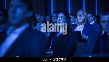 Porträt eines gutaussehenden Mannes, der in einem dunklen, überfüllten Auditorium auf einer Tech-Konferenz sitzt. Junger Mann konzentriert sich auf Keynote-Präsentation. Spezialist, inspiriert von den neuesten technologischen Fortschritten. Stockfoto