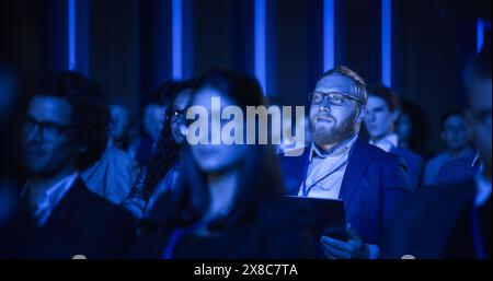 Porträt eines gutaussehenden Mannes, der in einem dunklen, überfüllten Auditorium auf einer Tech-Konferenz sitzt. Junger Mann konzentriert sich auf Keynote-Präsentation. Spezialist, inspiriert von den neuesten technologischen Fortschritten. Stockfoto