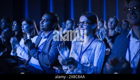 Junge Frau, die in einem überfüllten Publikum auf einer Geschäftskonferenz sitzt. Weibliche Teilnehmerinnen jubeln und klatschen nach einer Motivations-Keynote-Rede. Auditorium mit jungen erfolgreichen Geschäftsleuten. Stockfoto