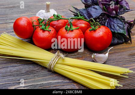 Vollkornspaghetti mit Zutaten auf einem Holztisch, Knoblauch, violettem Basilikum, frischen Tomaten auf einem Zweig Stockfoto