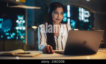 Indische Frau arbeitet am Abend in Creative Agency, Umsetzung moderner Geschäftsstrategie für Kunden. Erfolgreicher Spezialist, der einen Laptop für ernsthafte Arbeit verwendet. Stockfoto