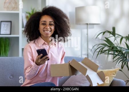 Im komfortablen Wohnzimmer packt eine Frau glücklich eine Lieferung auf einer grauen Couch aus, hält ihr Telefon, umgeben von Pflanzen, mit einem großen Lächeln und pflegt ein modisches, modernes Zuhause Stockfoto