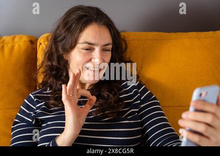 Eine Frau mittleren Alters lächelt und führt einen Videoanruf auf ihrem Smartphone durch, während sie das OK-Zeichen zeigt. Sie sieht glücklich und selbstbewusst aus, wenn sie mit ihr kommuniziert Stockfoto