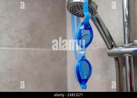 Eine blaue und gelbe Schwimmbrille hängt an einem Metallhaken in einem gut beleuchteten Duschraum, mit Wassertropfen an den Gläsern, bereit für Stockfoto