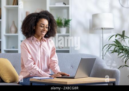Eine fröhliche Frau in Brille tippt auf einem Laptop auf einem Sofa in einem modernen Wohnzimmer mit Pflanzen und Bücherregalen im Hintergrund und schafft einen komfortablen und stilvollen Arbeitsplatz zu Hause Stockfoto
