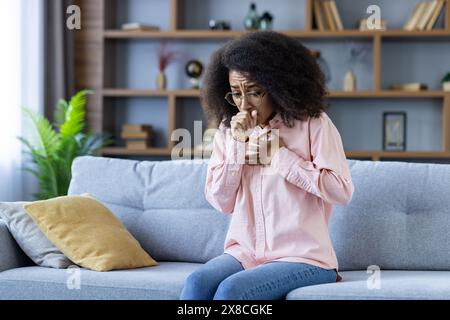 Eine Frau mittleren Alters mit Brille hustet in die Hand, während sie auf einer grauen Couch sitzt. Im Hintergrund befinden sich Regale mit Büchern und Pflanzen, die eine gemütliche Wohnzimmeratmosphäre schaffen. Stockfoto