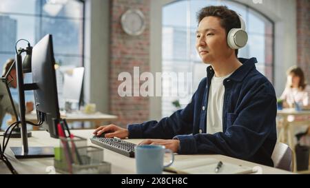 Porträt eines auf asiatische Kreationen fokussierten Mannes lächelt, während er Computer und Kopfhörer im hellen modernen Büro verwendet. Manager plant ein Projekt, während er sich Podcast anhört. Stockfoto