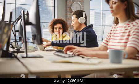 Gruppe von Kollegen, die im Büro an Computern arbeiten. Weibliche birassische Help Desk-Koordinatorin arbeitet mit asiatischen männlichen Kundendienstmitarbeitern mit Kopfhörnern, Stockfoto