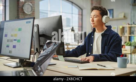Porträt eines asiatischen männlichen Mitarbeiters, der tagsüber am Computer in einem modernen Büro arbeitet. Der kreative Grafikdesigner inspiriert, während Sie Musik über Kopfhörer hören. Stockfoto