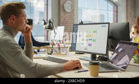 Porträt des weißen kreativen jungen Mannes mit Brille, der tagsüber in einem geräumigen modernen Büro an einem Computer arbeitet. Männlicher Grafikdesigner lächelt, während er ein neues visuelles Konzept für die Website entwickelt. Stockfoto