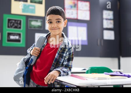 In der Schule steht ein kleiner birassischer Junge mit einem Rucksack lächelnd im Klassenzimmer, im Kopierraum Stockfoto