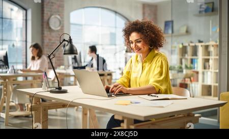 Porträt einer birassischen jungen Frau mit Laptop und Lächeln in einem geräumigen Büro. Technische Supportspezialistin für Frauen, die Probleme mit Software lösen. Kollegen, die im Hintergrund arbeiten. Stockfoto