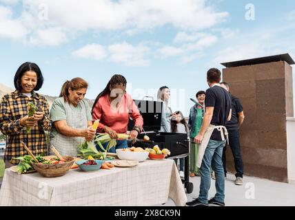 Generationenübergreifende Freunde, die Spaß beim Grillen auf dem Dach des Hauses haben - fröhliche, multirassische Menschen, die zusammen kochen Stockfoto