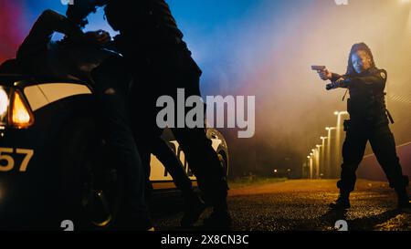 Männlicher Polizist zwingt einen gefährlichen Kriminellen auf die Haube des Polizeiautos und legt ihm Handschellen an. Kamerafokus n weiblich hält eine Waffe und Taschenlampe und zielt auf den Fellon. Stockfoto