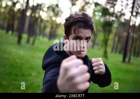 Sportlicher junger Mann in einer sportlichen schwarzen Jacke in einer Kampfposition, der seine Faust für einen Schlag vor einem Hintergrund von Waldbäumen hochhebt. Stockfoto