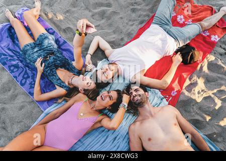 Gruppe von Freunden, die ein Selfie am Strand machen - Spaß und Entspannung genießen - auf Strandhandtüchern liegen, lächeln und lachen, Sommerurlaub und glücklich Stockfoto